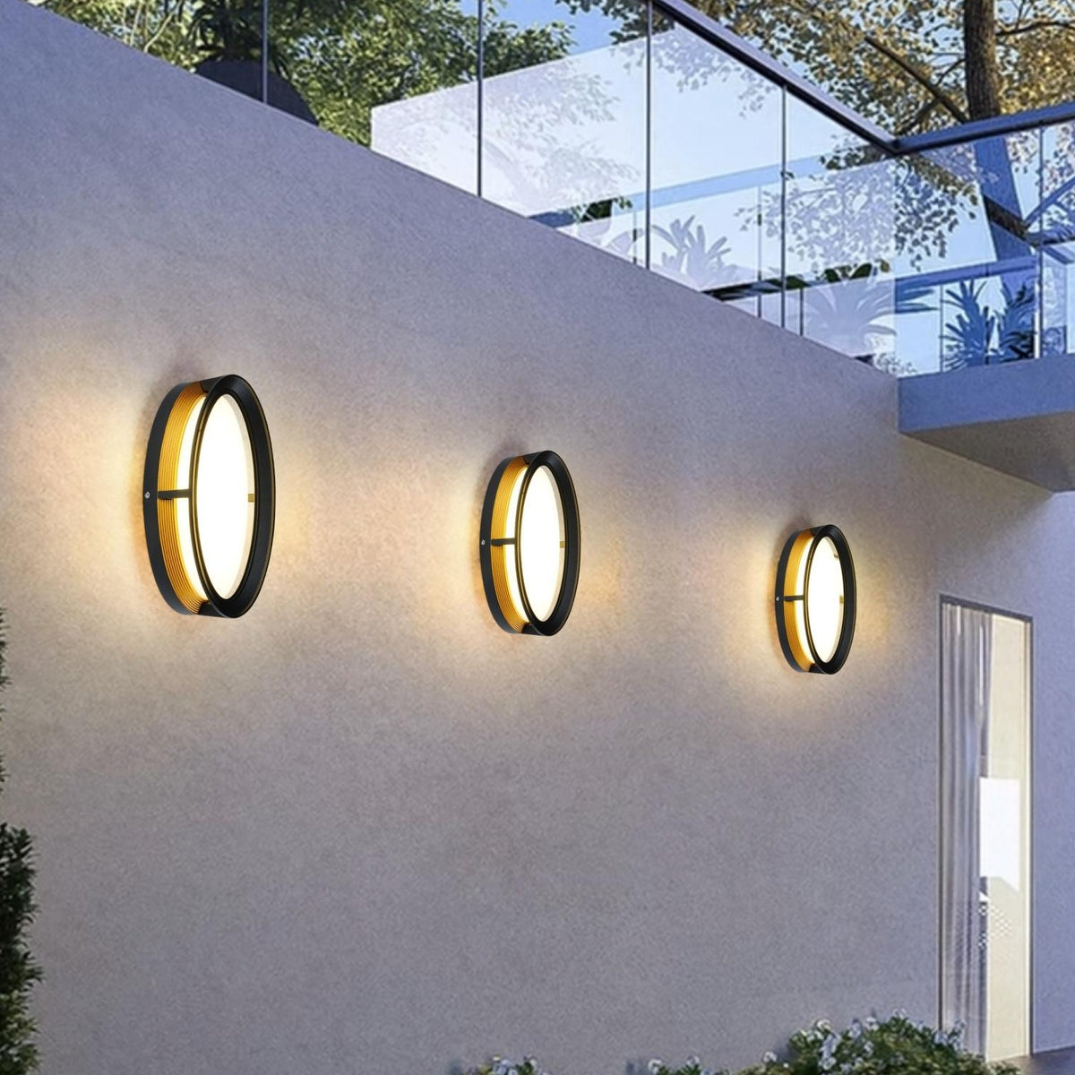 AURA Outdoor Wall Light – Round, Black & Gold (12W, Warm White 3000K) – lit on a garden wall at dusk