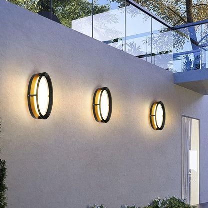 AURA Outdoor Wall Light – Round, Black & Gold (12W, Warm White 3000K) – lit on a garden wall at dusk